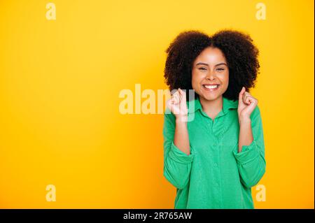 Aufgeregte frohe afroamerikanische oder brasilianische lockige Frau, Gesten mit den Fäusten, Freude am Erfolg, Erfolg, Erfolg, Sieg, Ein Lächeln vor der Kamera, isolierter gelber Hintergrund, Werbeschreibungsplatz Stockfoto