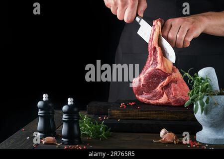 Der Koch schneidet Steak-Rindfleisch. Die Hände von Mans halten rohes Tomahawk-Steak auf rustikalem Holzbrett auf schwarzem Hintergrund. Kochen, Rezepte und Essenskonzept. S Stockfoto