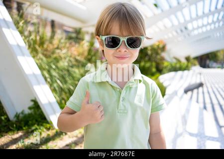 Nahaufnahme eines blonden kleinen Jungen mit Sonnenbrille und Daumen nach oben in einem Park im Freien Stockfoto