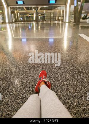 Mädchenbeine in roten Sneakers New Balance. Wartender Tourist im Abflugbereich am Flughafen. Allein am leeren Flughafen. Reisekonzept. Der letzte Passagier. Stockfoto