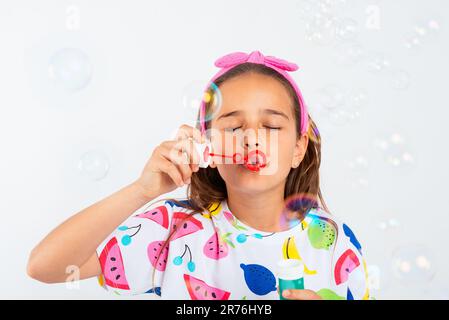 Das kleine Mädchen in bunten Kleidern bläst Seifenblasen, während es mit geschlossenen Augen vor weißem Hintergrund Spaß hat und beim Studiofoto spielt Stockfoto