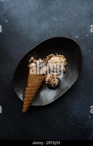 Draufsicht auf die schwarze Keramikplatte mit einer Schachtel Schokoladeneis, begleitet von zwei Kugeln Eiscreme mit Erdnüssen und Walnüssen Stockfoto