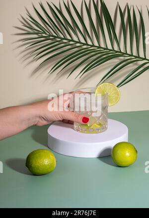 Anonyme Frau mit rotem Nagellack, Glas mit Cocktailgetränk mit Eis und Zitrusscheibe auf weißem Display auf dem Tisch mit Zitronen in der Nähe von Grün Stockfoto