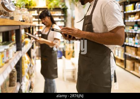 Gekürzter, nicht wiedererkennbarer afroamerikanischer Verkäufer in einer Schürze, der neben Regalen steht, Lebensmittel einkauft und auf den Bildschirm eines Tablets schaut, während er nachforscht Stockfoto