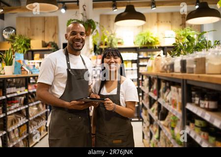 Fröhliche junge afroamerikanische Verkäufer in Schürzen stehen zwischen Schränken mit Lebensmitteln, halten ein Tablet und lächeln in die Kamera Stockfoto