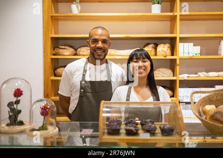 Positive junge, multiethnische Kollegen in Schürzen, die lächeln und in die Kamera schauen, während sie während der Arbeitszeit hinter dem Tresen der Bäckerei stehen Stockfoto