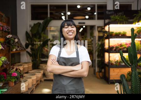 Glückliche junge asiatische Floristin in der Schürze, die lächelt und in die Kamera schaut, während sie in einem Blumenladen mit gekreuzten Armen steht Stockfoto