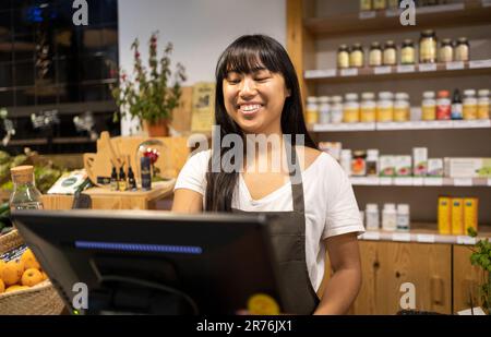 Glückliche junge asiatische Verkäuferin in der Schürze lächelt und verwendet Computer, während sie an der Theke mit verschiedenen Produkten während der Arbeit im Supermarkt steht Stockfoto