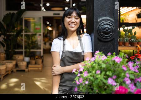 Fröhliche junge asiatische Floristin in der Schürze, die lächelt und in die Kamera schaut, während sie neben dem Tresen mit blühenden Blumen während der Arbeit in Floristik Sho steht Stockfoto