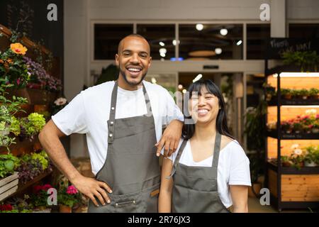 Fröhliche junge, multiethnische Kollegen in Schürzen, die lächeln und vor die Kamera schauen, während sie während des Arbeitsprozesses im Blumenladen zusammen stehen Stockfoto