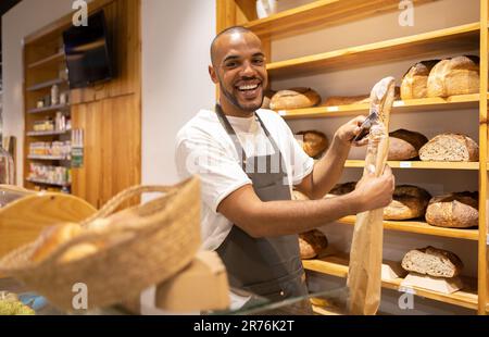 Glücklicher junger afroamerikanischer Verkäufer in der Schürze, der frisch gebackenes Brot in eine Papiertüte legte, während er während des Arbeitsvorgangs neben Regalen stand Stockfoto