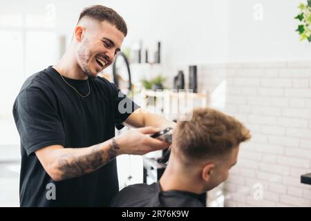 Seitenansicht eines jungen Barbiers mit Tätowierungen, die unerkannte Männerhaare mit Haarschneidern vor verschwommenem Hintergrund schneiden Stockfoto