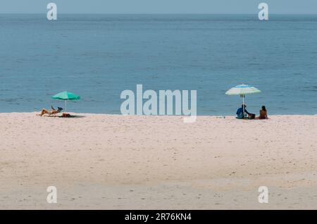 Eine Gruppe von Menschen, die einen sonnigen Tag am Strand genießen, sich unter bunten Sonnenschirmen entspannen und den Blick auf das türkisfarbene Meer genießen Stockfoto