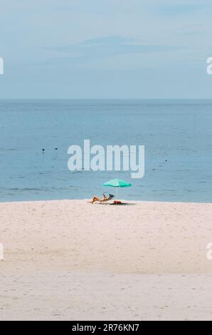 Eine junge Frau, die sich an einem sonnigen Strandtag auf einem Stuhl entspannt, geschützt durch einen Sonnenschirm für Schatten Stockfoto