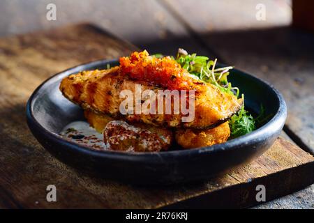 Mit Sauce und Kräutern garniertes gegrilltes Lachssteak, serviert auf einer schwarzen Schüssel auf einem Holzbrett Stockfoto