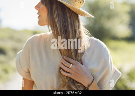 Porträt einer lächelnden jungen Frau mit Strohhut, die den Hinterkopf mit den Fingern berührt und bei Sonnenlicht vor verschwommenem Hintergrund wegschaut Stockfoto