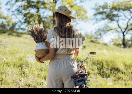 Rückblick auf ein nicht wiedererkennbares Weibchen mit Strohhut, das Lavendel- und Weizenstrauß trägt und am sonnigen Tag auf dem Land mit dem Fahrrad steht Stockfoto