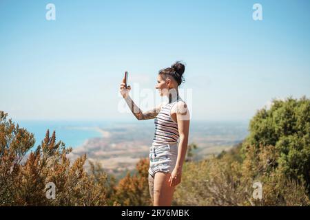 Fröhliche junge Frau in lässiger Kleidung mit stilvoller Frisur und Tattoo, die Selfie auf dem Smartphone macht, während sie auf dem Land steht Stockfoto