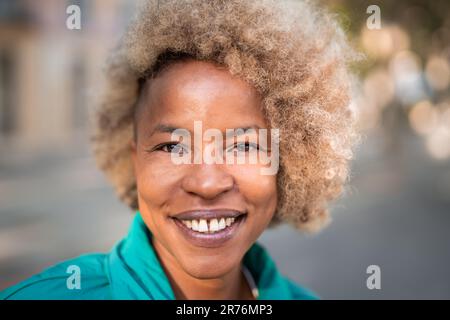 Porträt einer fröhlichen afroamerikanischen Frau mit lockigem, lächelndem Haar und Blick in die Kamera, während sie auf der Straße mit verschwommenem Hintergrund steht Stockfoto
