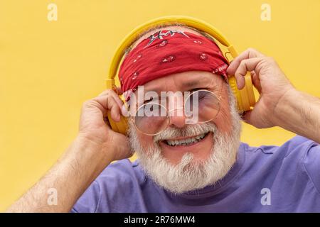 Fröhlicher Hipster Senior mit Bart und Kopfhörer, der Musik hört und mit einer Sonnenbrille in die Kamera schaut, während er die Kopfhörer gegen einen hält Stockfoto