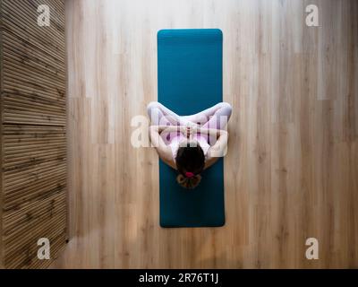 Blick von oben auf eine Frau in pinkfarbener Sportbekleidung, die in Lotusposition auf einer Yogamatte sitzt und meditiert, betet oder Achtsamkeit übt. Mit ihr Stockfoto