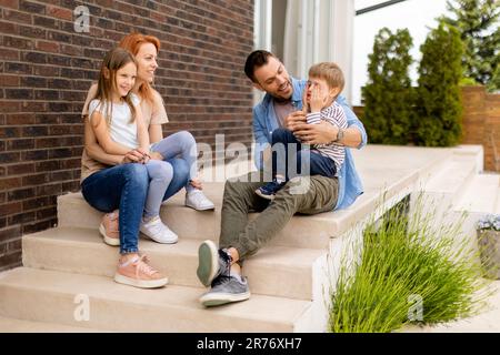 Familie mit einer Mutter, einem Vater, einem Sohn und einer Tochter, die draußen auf den Stufen einer Veranda eines Backsteinhauses sitzen Stockfoto