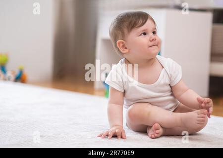 Ein dicker kleiner Junge im weißen Bodysuit, der auf einem flauschigen Teppich sitzt, der im Kinderzimmer auf dem Boden liegt Stockfoto