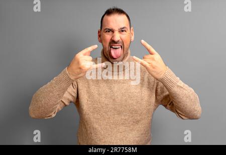 Die Show muss weitergehen. Foto eines Mannes, der seine Zunge aus dem Mund steckte und Hörner machte, Rock-and-Roll-Lover in beigefarbenem Rollkragenpulli, isoliert über Stockfoto