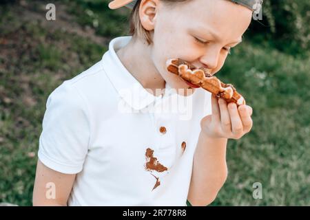 Schokoladenflecken auf weißen Kleidern. Ein Junge isst belgische Waffeln mit klebrigen Schokoladenhänden. Verdorbene Kleidung. Im Freien. Stockfoto