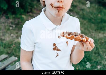 Schokoladenflecken auf weißen Kleidern. Ein Junge isst belgische Waffeln mit klebrigen Schokoladenhänden. Verdorbene Kleidung. Im Freien Stockfoto
