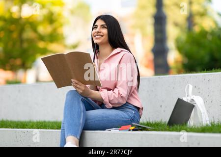 Ziemlich glückliche hispanische Dame, die einen Stift in einem Notizbuch hält, mit einem Laptop im Park sitzt, denkt, plant die Zukunft Stockfoto