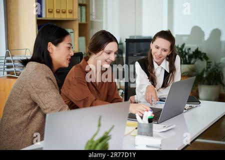 Gruppe von Kollegen, die Online-Präsentationen auf einem Laptop diskutieren, während sie zusammen am Tisch sitzen und ein Notebook im Büro verwenden Stockfoto