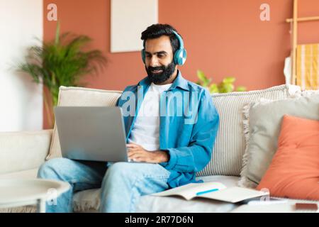 Glücklicher Arabier, Der Ein Notebook Mit Kopfhörern Benutzt Und Von Zu Hause Aus Arbeitet Stockfoto