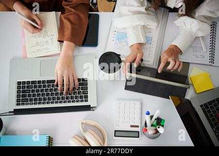 Draufsicht von Geschäftsfrauen, die im Team arbeiten, über Projekte, die mit Geräten und Dokumenten am Tisch sitzen Stockfoto