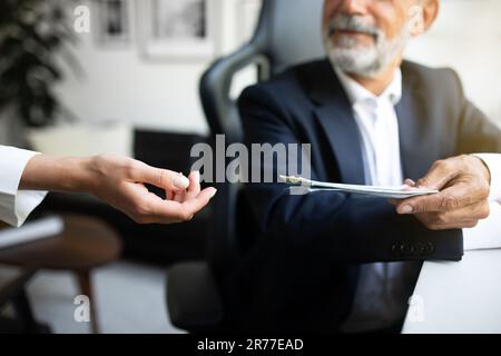 Lächelnder europäischer Geschäftsmann im Anzug, der am Tisch sitzt, spendet der Frau Geld Hand in Amt Stockfoto