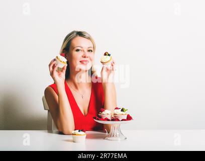 Wunderschöne junge blonde Frau, die am Tisch sitzt und selbstgemachte Cupcakes hält Stockfoto