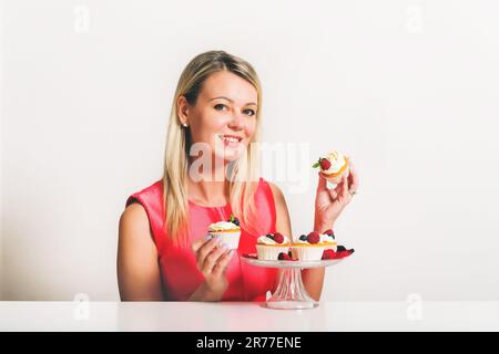 Wunderschöne junge blonde Frau, die am Tisch sitzt und selbstgemachte Cupcakes hält Stockfoto