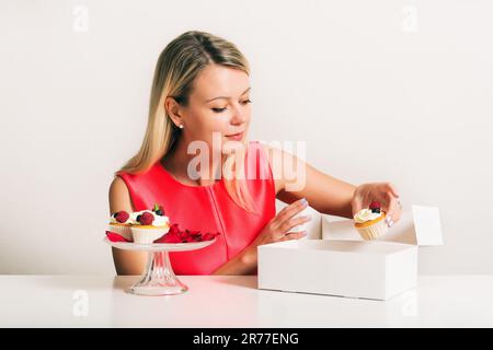 Wunderschöne junge blonde Frau, die frische hausgemachte Cupcakes in eine weiße Schachtel packt Stockfoto