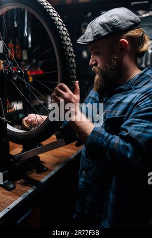 Vertikale Seitenansicht eines bärtigen Fahrradmechanikers, der die Fahrradspeiche mit einem Radspeichenschlüssel in einer Fahrradwerkstatt mit dunklem Innenraum überprüft. Stockfoto