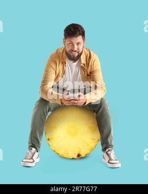 Junger Mann mit Bart und Handy, der auf einem großen Stück Ananas vor blauem Hintergrund sitzt Stockfoto