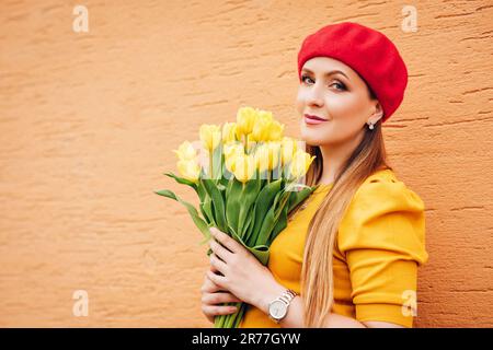 Ein Frühlingsporträt von einer jungen, wunderschönen Frau im Freien, die ein gelbes Oberteil und eine rote Baskenmütze trägt, einen Strauß frischer Tulpenblüten hält und gegen die orangefarbene Wand posiert Stockfoto