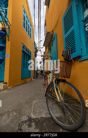 Ein altes Fahrrad steht in der engen Straße, umgeben von gelben Gebäuden mit blauen Fenstern und Türen. Altstadt von Hoi an, Vietnam. Stockfoto
