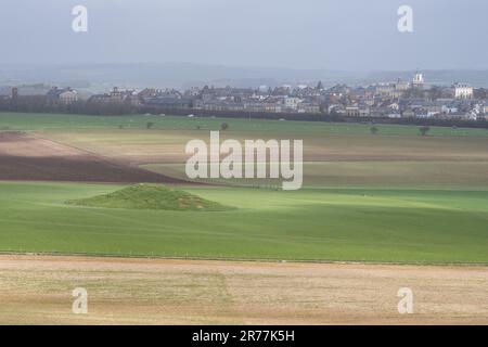 Ein alter Tumulus-Grabhügel steht auf einem Feld unterhalb von Maiden Castle in Dorset, mit Poundbury Neustadt in der Ferne. Stockfoto