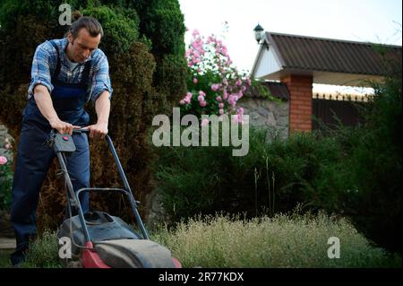 Hispanischer Gärtner in kariertem blauen Hemd und Arbeitsgartenuniform mäht den Rasen mit einem elektrischen Rasenmäher im Garten Stockfoto