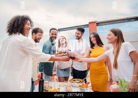 Gruppe von erwachsenen Freunden, die auf einer Dinnerparty auf dem Dach lächeln und ein Glas Wein trinken. Junge glückliche Menschen, die Spaß haben, mit Alkohol anzustoßen, feiern zusammen auf einem freundlichen Treffen. Hochwertiges Foto Stockfoto