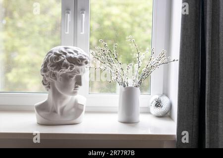 Apollos Gipskopf in einem weißen Innenraum. Eine Spiegeldisco-Kugel und verwelkte Weidenzweige in einem weißen Krug auf der Fensterbank neben dem Fenster schmücken das Stockfoto