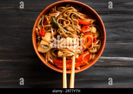 Teller mit Buchweizennudeln mit Gemüse, Pilzen, Hühnerfleisch auf dunklem Hintergrund, Draufsicht. Chinesische Stöcke nehmen japanische Soba von einem Clay bo Stockfoto