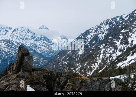 Niedrige Wolken hängen über schneebedeckten Bergen mit Flechten bedeckt und großen Steinen im Vordergrund Stockfoto