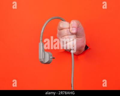 Die Hand eines Mannes hält ein graues Stromkabel durch gerissenes orangefarbenes Papier. Hand durch zerrissenes orangefarbenes Papier. Stockfoto