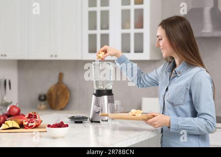 Wunderschöne junge Frau, die Banane in den Mixer gibt, um leckeren Smoothie in der Küche zu erhalten Stockfoto